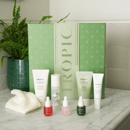 The Advanced Glow Facial - active skincare collection by Tropic. A large green festive gift set box with a full routine displayed in front: Smoothing Cleanser, Hydrating Cleanser, Skin Feast moisturiser, Juicy Greens essence, and three serum drops — Glow Berry, Fruit Peel, and Super Greens.